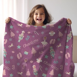 May include: A purple blanket featuring a pattern of ballet-themed illustrations, including ballerinas, nutcrackers, and Christmas trees. The name "Charlotte" is printed on the blanket in white. The blanket is made of a soft material.