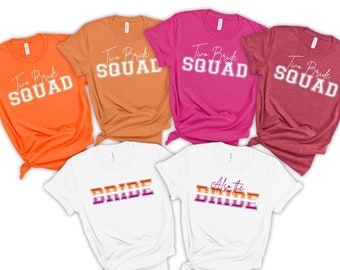May include: Five t-shirts with different designs for a wedding party. Two shirts say "Two Bride Squad" in white script on orange, rust, and maroon shirts. Two white shirts say "Bride" and "Also the Bride" in rainbow colors.
