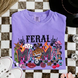 May include: A lilac Comfort Colors t-shirt featuring the text "FERAL MAMA" in a graphic design. The design incorporates a tiger, playing cards, a skull, and other colourful elements. The shirt is displayed on a checkered surface.