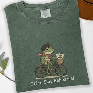 May include: Green t-shirt with a frog riding a bicycle, wearing a beret and scarf, and holding a script. The bicycle has a basket with theater masks. Text at the bottom reads "Off to Slay Rehearsal."