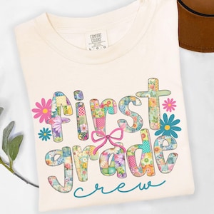 May include: A cream-coloured t-shirt featuring the text "First Grade Crew" in patchwork-style letters with floral patterns and a pink bow. The shirt is folded, with a tag visible. The design is colourful.