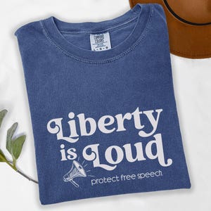 May include: A navy blue t-shirt featuring the white text "Liberty is Loud", a megaphone graphic, and the words "protect free speech". The shirt is folded and has a Comfort Colors label.