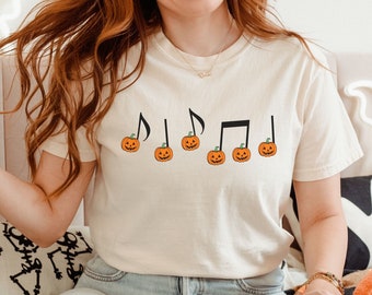 May include: A white t-shirt with a black musical note design and orange pumpkins. The pumpkins have green stems and are arranged in a line.