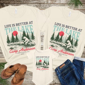 K&ouml;nnte beinhalten: Zwei cremefarbene T-Shirts und ein cremefarbener Baby-Body, jeweils mit einer Grafik eines b&auml;ren, der an einem See mit Tannen und einer roten Sonne angelt. Der Text lautet: LIFE IS BETTER AT THE LAKE, Camp Anderson, LAKE TAHOE, CA 2025.