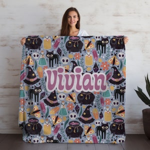 May include: A soft, blue fleece blanket with a whimsical Halloween design featuring black cats, cauldrons, potion bottles, and crystals. The name "Vivian" is written in pink across the center of the blanket.