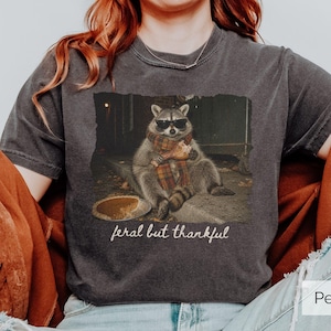 May include: A charcoal gray t-shirt featuring a graphic of a raccoon wearing sunglasses and a plaid scarf, holding a turkey leg next to a pumpkin pie. The text below the raccoon reads feral but thankful. The shirt has a vintage washed finish.
