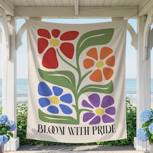 May include: A cream-colored tapestry with a floral design featuring red, orange, blue, and purple flowers with green leaves. The text "BLOOM WITH PRIDE" is printed in black at the bottom of the tapestry. The tapestry is hanging on a porch overlooking the ocean.