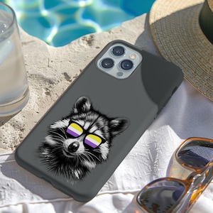 May include: A dark gray phone case featuring a raccoon wearing non-binary pride flag sunglasses. The case is resting on a white surface next to a pair of sunglasses and a glass of water. A straw hat is in the background.