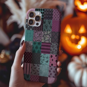 May include: A patchwork phone case in shades of teal, purple, and gray, featuring a variety of patterns including skulls, bats, and stripes. The case is held in a hand, with a blurred background of pumpkins and fall decor.
