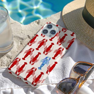 May include: A white iPhone case with a pattern of red lobsters and one blue lobster. The case is lying on a white surface with a straw hat and sunglasses nearby.