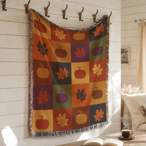 May include: A woven throw blanket with a patchwork design featuring pumpkins and maple leaves in shades of orange, red, green, and purple. The blanket is rectangular with a fringed edge and is hanging on a white wall.