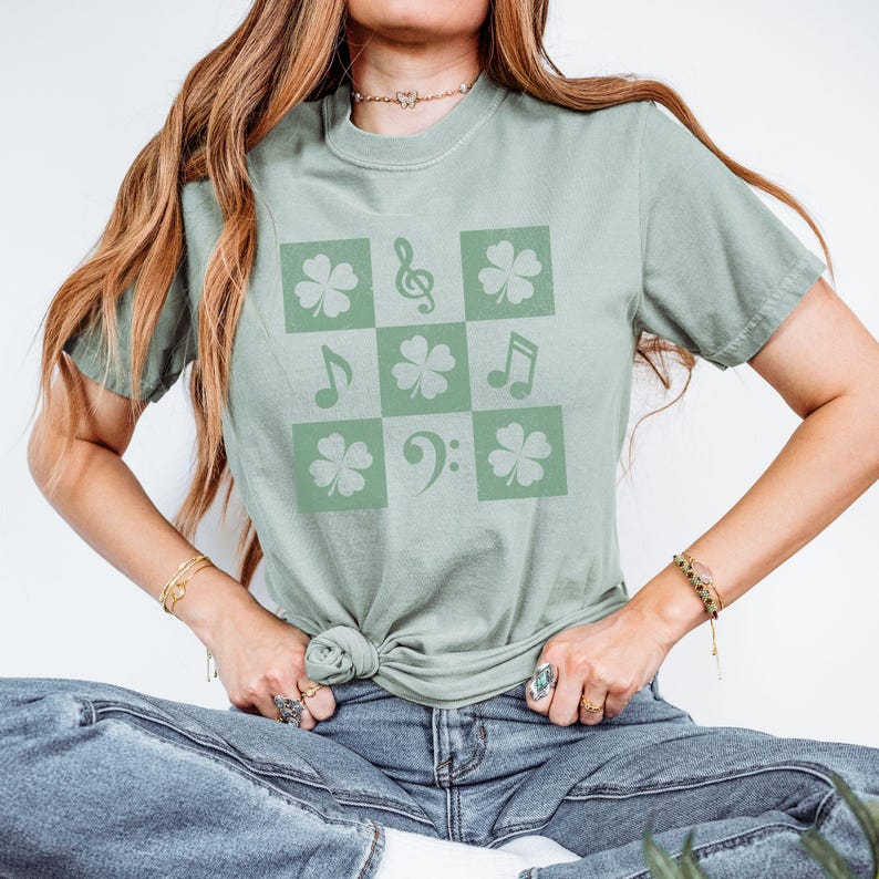 May include: A sage green t-shirt with a graphic design of a checkered pattern featuring shamrocks and musical notes.