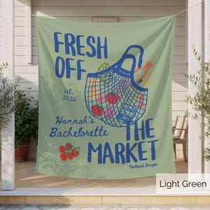 Peut inclure: Une banni&egrave;re en tissu vert clair avec un graphique bleu repr&eacute;sentant un sac de march&eacute; en filet rempli de l&eacute;gumes. Le texte indique FRESH OFF THE MARKET, est. 2026, Hannah's Bachelorette, et Portland, Oregon, avec des tomates rouges dessin&eacute;es.