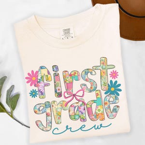 May include: A cream-colored t-shirt featuring the text First grade in a multi-colored floral and checkered patchwork pattern. A pink bow graphic sits between the words, accompanied by pink and blue flower icons and the word crew in teal.