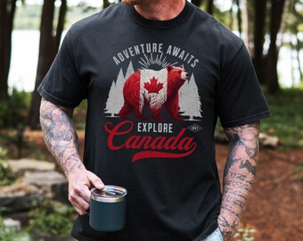 May include: A black short-sleeve t-shirt featuring a graphic of a bear with the Canadian flag pattern on its fur, surrounded by pine trees. The text reads ADVENTURE AWAITS and EXPLORE Canada with the year 1867 in a small diamond shape.