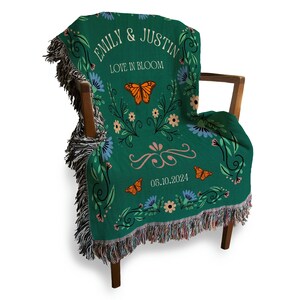 May include: A green woven throw blanket with a floral design and butterflies. The blanket has the text "Emily & Justin Love in Bloom 05.10.2024" in white.