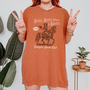 May include: A rust-colored t-shirt with a graphic of a cowgirl riding a horse and reading a book. The text on the shirt reads "Books, Boots & Broncs Cowgirl Book Club".