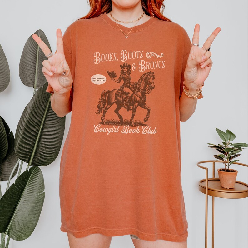 May include: A rust-colored t-shirt with a graphic of a cowgirl riding a horse and reading a book. The text on the shirt reads "Books, Boots & Broncs Cowgirl Book Club".