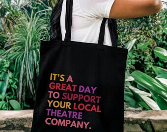 May include: A black tote bag with two shoulder straps featuring the multi-colored text IT'S A GREAT DAY TO SUPPORT YOUR LOCAL THEATRE COMPANY. The lettering is printed in shades of pink, orange, yellow, and purple against the black fabric.