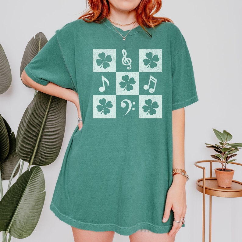 May include: A teal green t-shirt with a white graphic of a checkered pattern with shamrocks, musical notes, and a bass clef.