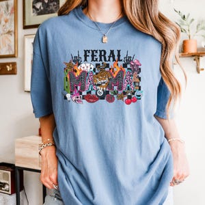 May include: A pale blue t-shirt featuring the words "FERAL MAMA" in a vibrant graphic. The design incorporates a tiger, flames, and various other motifs. The shirt is made from a soft fabric.