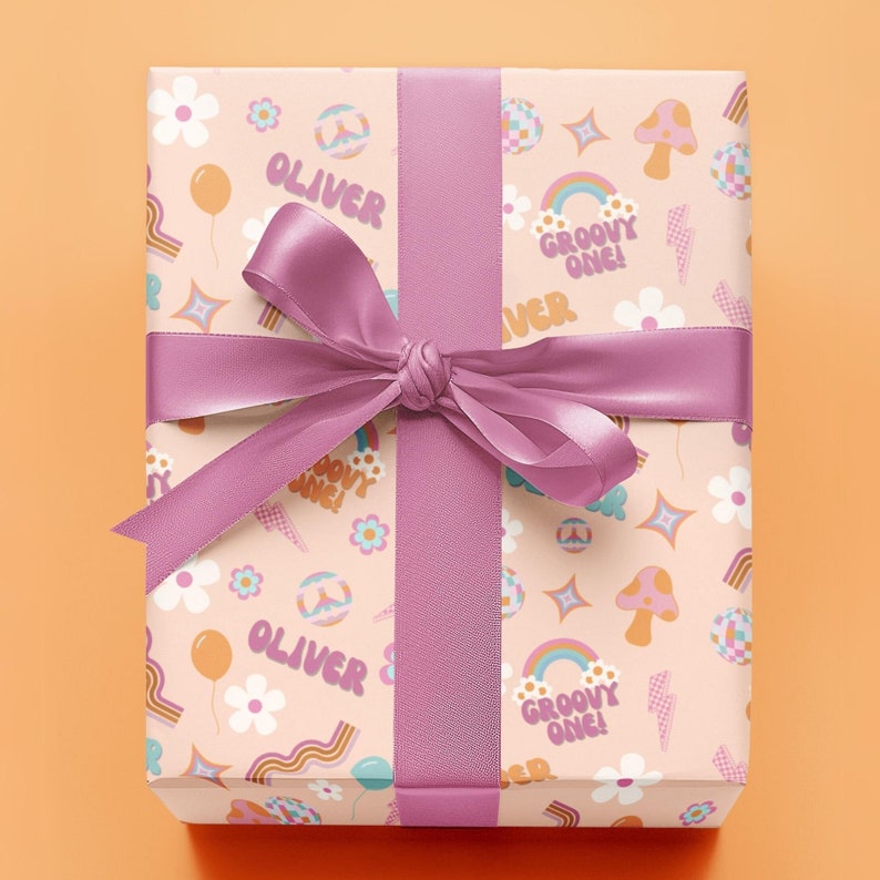 May include: A pink gift box with a pink ribbon and a groovy 70s themed wrapping paper. The wrapping paper features a pattern of peace signs, rainbows, mushrooms, and the text "Oliver" and "Groovy One!"