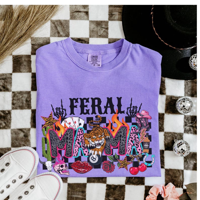May include: A lilac Comfort Colors t-shirt featuring the text "FERAL MAMA" in a graphic design. The design incorporates a tiger, playing cards, a skull, and other colourful elements. The shirt is displayed on a checkered surface.