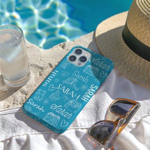May include: A teal phone case with the name "Sarah" in white script and repeating line art of books, flowers, and other objects. The phone case is next to sunglasses, a straw hat, and a glass of water.
