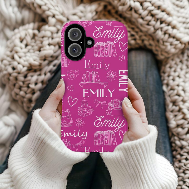 May include: A magenta phone case with a white pattern of books, hearts, flowers, and the name "Emily" repeated throughout. The case is held in a person's hands.
