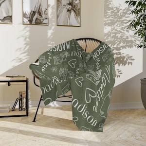 May include: A green throw blanket with a white design featuring the name "Madison" repeated throughout. The blanket is draped over a wicker chair.