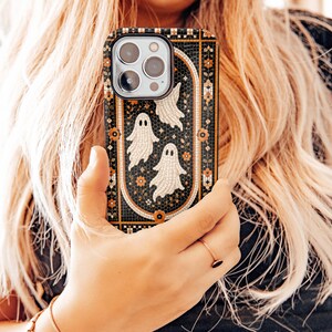 May include: A rectangular smartphone case featuring a black mosaic tile background with an arched border. The design includes three white ghost figures and small orange floral accents arranged within the central arch, creating a detailed pattern.