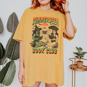 Pode incluir: Camiseta amarelo mostarda com um gr&aacute;fico que diz "Summerween Book Club". O gr&aacute;fico apresenta uma bruxa, um fantasma e uma mulher sob um guarda-chuva, todos lendo livros. Um gato preto est&aacute; sentado perto da bruxa. A camisa tem um ajuste folgado.