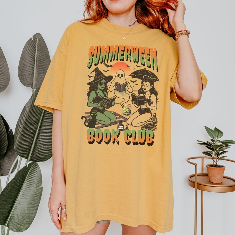 Pode incluir: Camiseta amarelo mostarda com um gr&aacute;fico que diz "Summerween Book Club". O gr&aacute;fico apresenta uma bruxa, um fantasma e uma mulher sob um guarda-chuva, todos lendo livros. Um gato preto est&aacute; sentado perto da bruxa. A camisa tem um ajuste folgado.