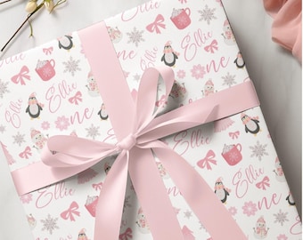 May include: A square gift box wrapped in white paper featuring a repeating pattern of pink bows, snowflakes, penguins, snowmen, and mugs, with the name Ellie and the word one printed in pink script, tied with a wide pink satin ribbon.
