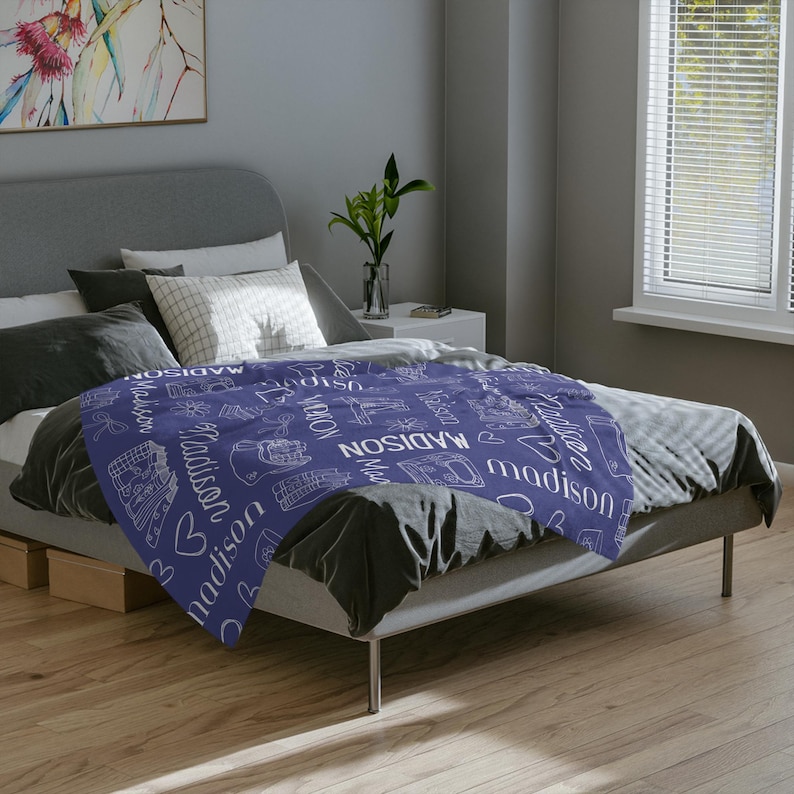 May include: A purple throw blanket with a white pattern of the name "Madison" repeated throughout. The blanket is on a bed with white pillows.