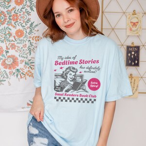 以下が含まれることがあります： 薄い青色のTシャツに、ベッドで本を読んでいる女性のヴィンテージスタイルのイラストが描かれています。Tシャツのテキストには「My idea of Bedtime Stories has definitely evolved!」と、赤い星評価と赤い円の中に「Extra Spicy!」と書かれています。「Smut Readers Book Club」というテキストはイラストの下に印刷されています。
