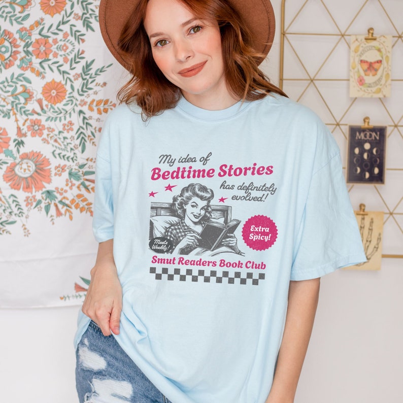 以下が含まれることがあります： 薄い青色のTシャツに、ベッドで本を読んでいる女性のヴィンテージスタイルのイラストが描かれています。Tシャツのテキストには「My idea of Bedtime Stories has definitely evolved!」と、赤い星評価と赤い円の中に「Extra Spicy!」と書かれています。「Smut Readers Book Club」というテキストはイラストの下に印刷されています。