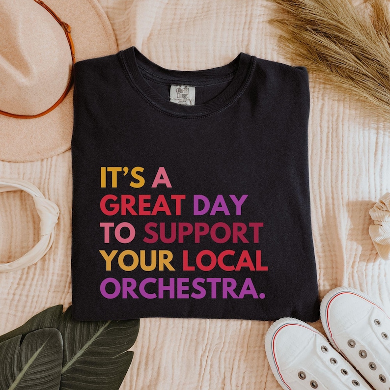 May include: Black t-shirt with the text "It's a great day to support your local orchestra." in rainbow colors.