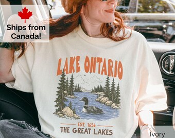 May include: An ivory t-shirt featuring a graphic print of a loon swimming in a lake surrounded by pine trees. The text reads Lake Ontario at the top and Est. 1656 The Great Lakes at the bottom in orange and brown lettering.