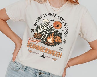 May include: A cream-colored t-shirt featuring a graphic of a skeleton sitting by a campfire with a tent, a full moon with a witch silhouette, and the text "WHERE SUMMER GETS SPOOKY!", "Camp SUMMERWEEN", and "Since 1976" with broomstick icons.