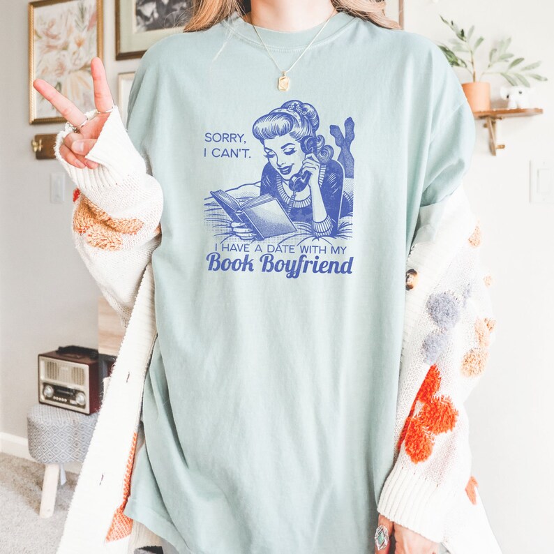 Op de afbeelding: Een lichtblauw T-shirt met een vintage illustratie van een vrouw die een boek leest en telefoneert. De tekst op het shirt luidt "Sorry, I can't. I have a date with my Book Boyfriend."