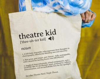May include: A cream-colored canvas tote bag featuring black printed text that defines the term theatre kid. The design includes a phonetic pronunciation guide, a speaker icon, and three numbered descriptive points about the noun.