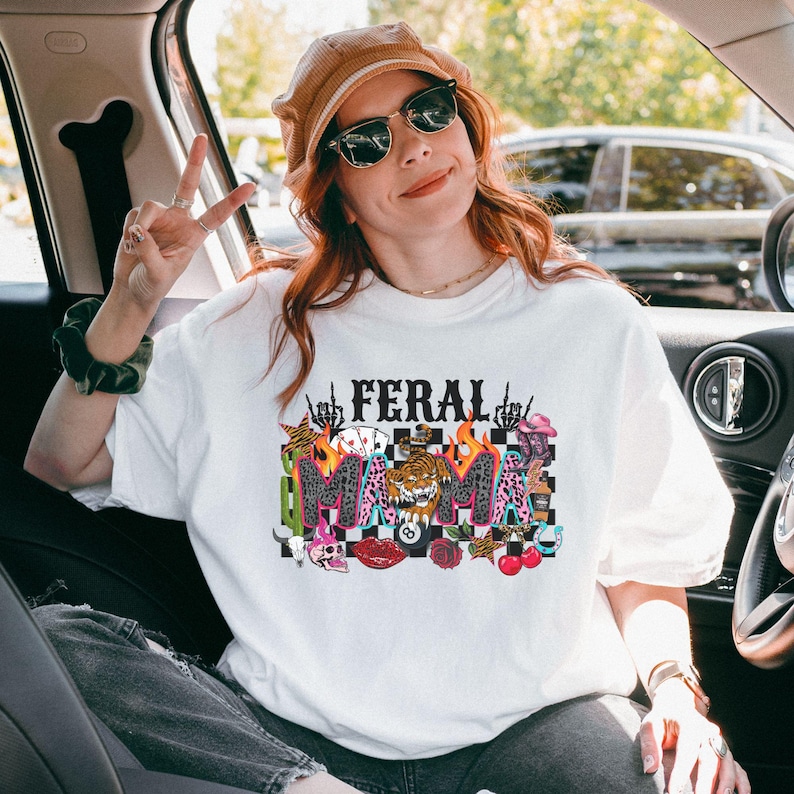 May include: White t-shirt featuring the text "FERAL MAMA" in a vibrant graphic. The design incorporates flames, a tiger, playing cards, and other illustrations. The shirt is a relaxed fit.