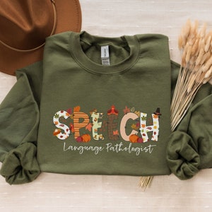 Peut inclure: Un sweat-shirt à manches longues vert avec un motif sur le thème de l'automne qui dit "SPEECH Language Pathologist". Les lettres sont décorées de feuilles d'automne, de citrouilles et d'autres images d'automne.
