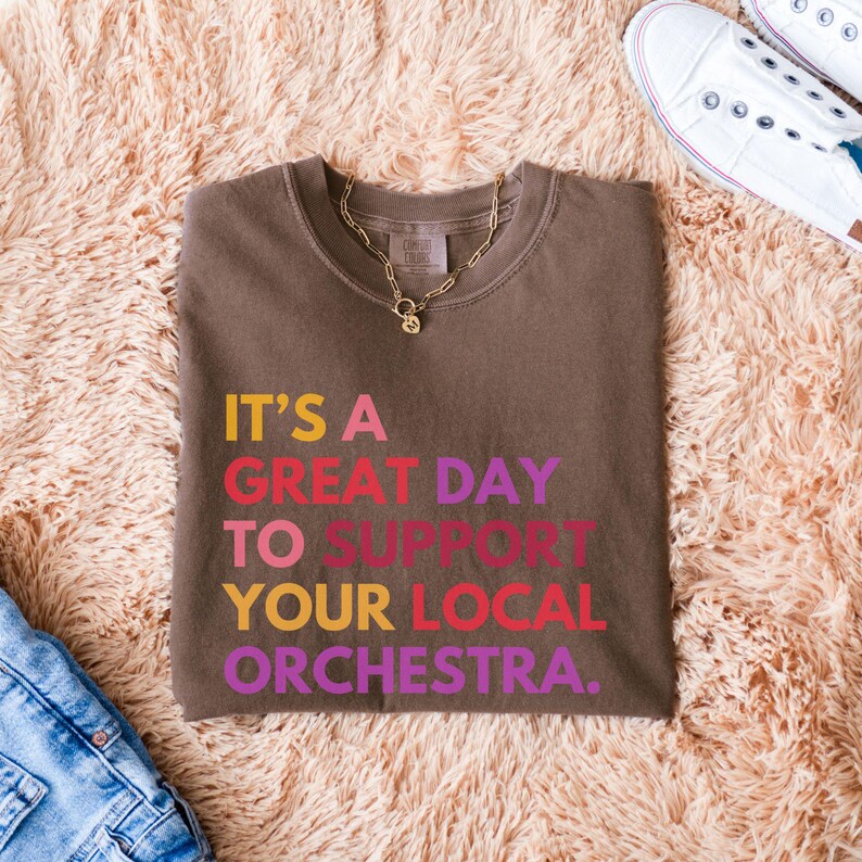 May include: A brown t-shirt with the text "It's a great day to support your local orchestra." printed in rainbow colors.
