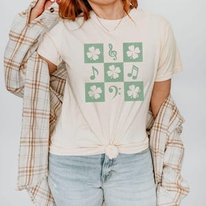 May include: A cream colored t-shirt with a green shamrock and musical note design. The design is in a checkered pattern.