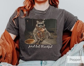 May include: A charcoal gray short-sleeve t-shirt featuring a graphic of a raccoon wearing sunglasses and a plaid scarf, holding a turkey leg next to a pie. The text below the raccoon reads feral but thankful. The shirt has a crew neckline.