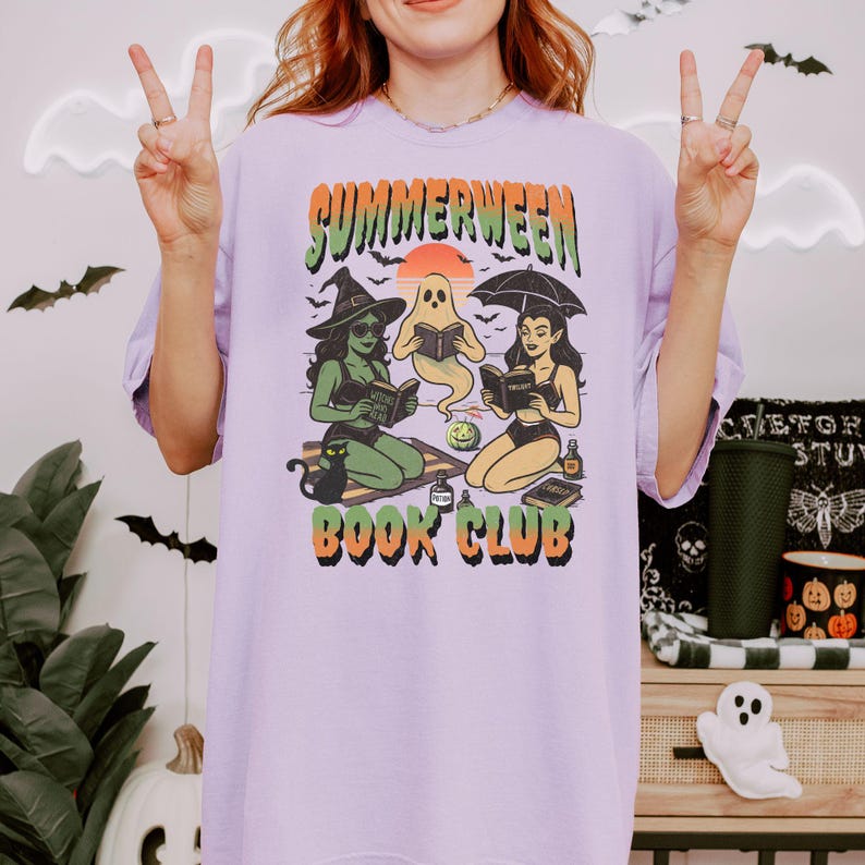 Pode incluir: Camiseta lil&aacute;s clara com a frase "SUMMERWEEN BOOK CLUB" em laranja e verde. O desenho mostra uma bruxa, um fantasma e uma mulher lendo livros. O fundo inclui morcegos e um p&ocirc;r do sol. Design caprichoso.