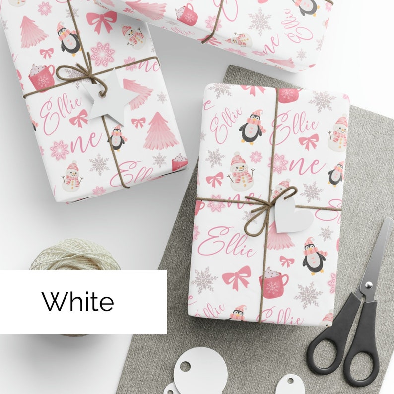 May include: White wrapping paper with a pink and silver snowflake pattern. The paper features a penguin, a snowman, a hot chocolate mug, and the name "Ellie" repeated throughout the design.