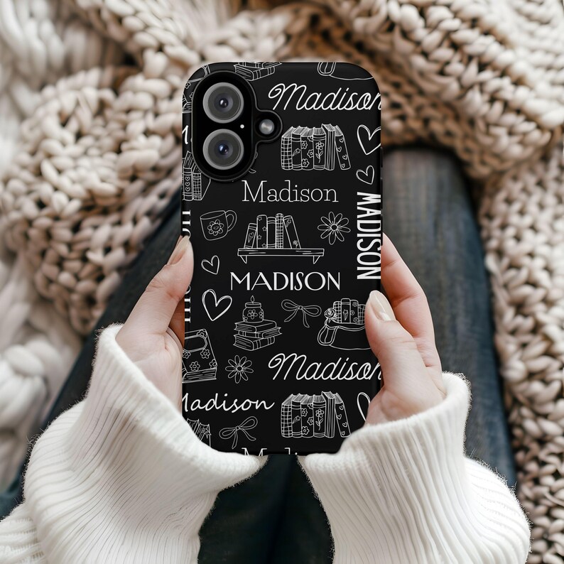 May include: Black phone case with a white doodle pattern of books, hearts, flowers, and the name "Madison" in a handwritten font. The phone case is held in front of a cozy, neutral-toned background.